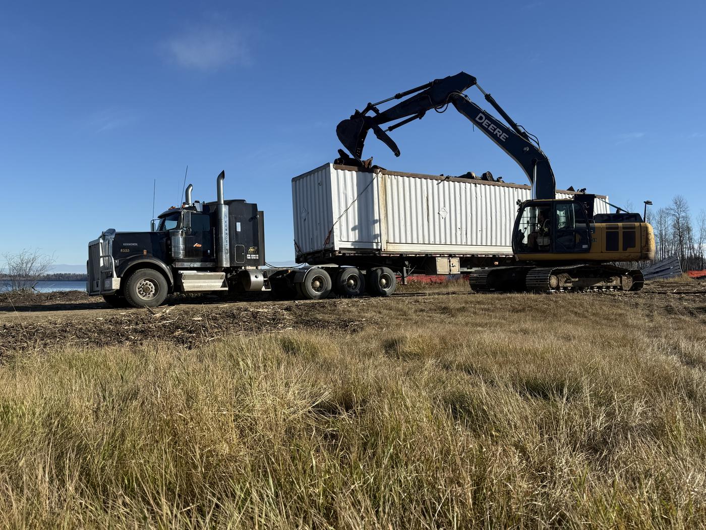 Excavator loading recovered metal into a shipping container on a Kenworth trailer at a lease site